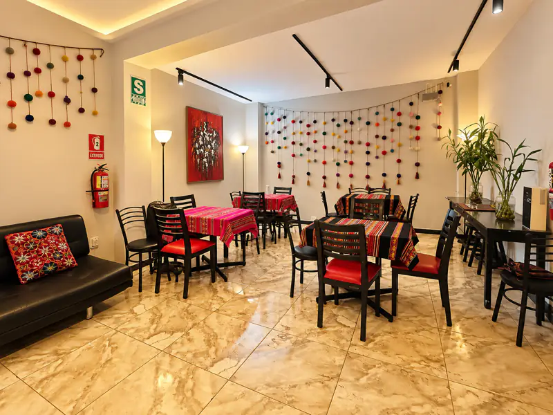 Vista completa de la sala de eventos en Lince: cuatro mesas con manteles andinos, sillas rojas y negras, sofá de cuero y plantas junto a la pared de pompones